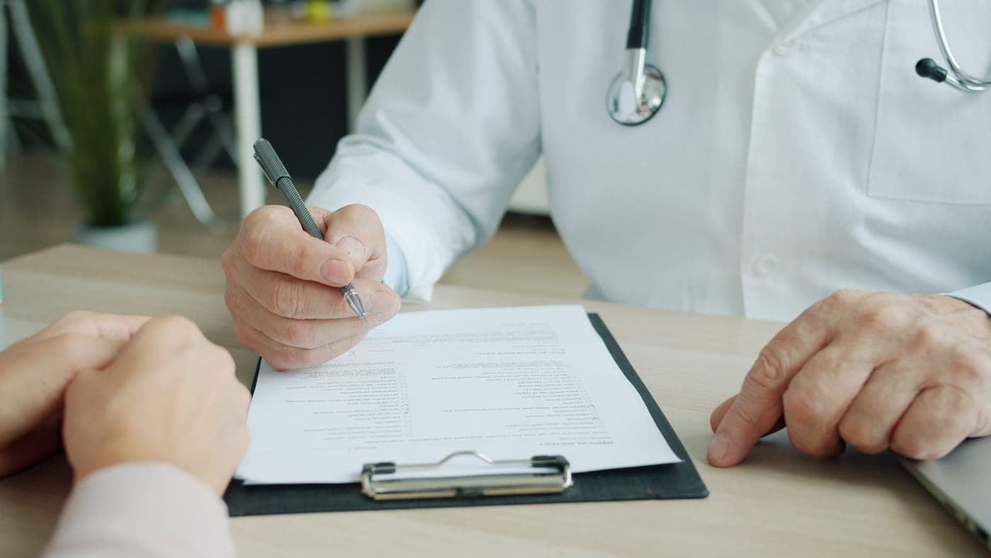 Médico com estetoscópio preenchendo um documento ao lado do paciente durante a consulta